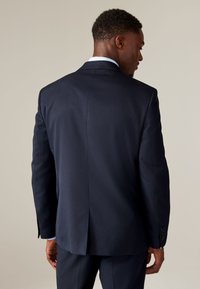 Tumesinine ülikonnajakk tekstureeritud kangaga, struktureeritud õlgadega ja ühe tagasõõrmega. Mansetitel nupud, seljal puhas disain ja nähtav hele vooder.