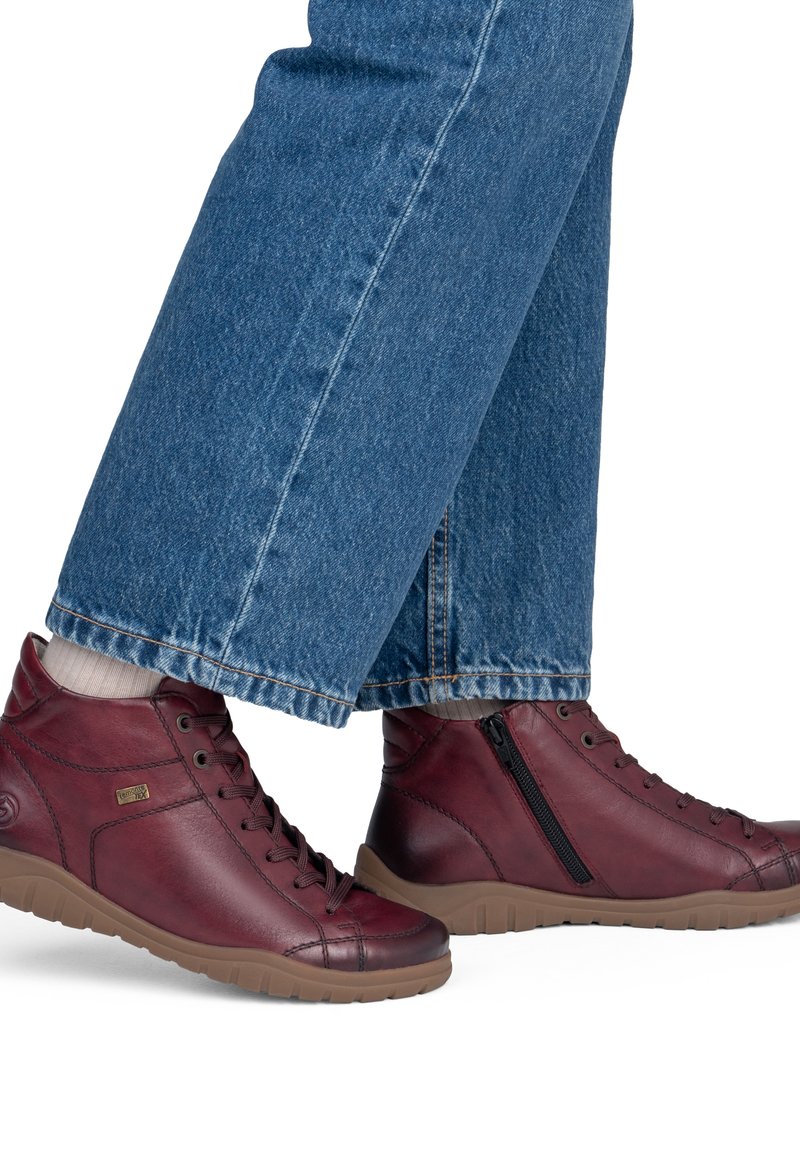 Personne portant un jean bleu et des baskets montantes en cuir bordeaux avec des semelles beiges et des fermetures éclair, debout sur un fond blanc.