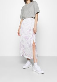 Osoba ubrana w szary t-shirt Nike, spódnicę w białe i fioletowe marmurkowe wzory z przednim rozcięciem, białe skarpetki i białe trampki, stojąca na jednolitym tle.