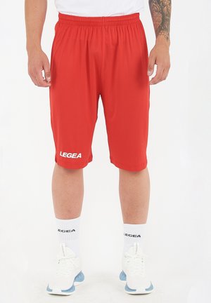 Uomo che indossa pantaloncini sportivi rossi con il logo "LEGEA", calzini bianchi e scarpe da ginnastica bianche, in piedi davanti a uno sfondo bianco semplice.
