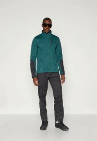 Mann med svarte solbriller, teal zip-up jakke med svarte ermer, svarte bukser og svarte sneakers, som står mot en enkel hvit bakgrunn.