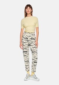 Cremefarbenes, geripptes Kurzarmoberteil, gemusterte beige Hose mit schwarzen Streifen und silberne Turnschuhe mit gelben Socken. Schlichtes Design, lockere Passform.