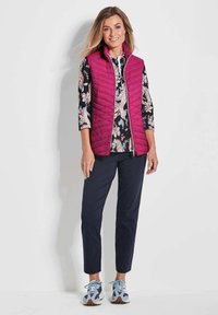 Gequiltte vest in levendig fuchsia, gebloemd shirt met florale ontwerpen, donkerblauwe broek en lichtblauwe sneakers met gemixte accenten.