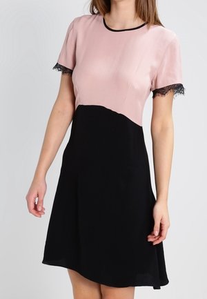 Kvinde iført en kortærmet kjole med en pink top, sort nederdel og sort blonder trim på ærmekanter, står mod en ensfarvet baggrund.