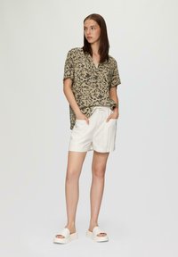 Lässiges, kurzärmliges Hemd in Beige mit schwarzem Blumenmuster, kombiniert mit weißen Leinen-Shorts und Plateausandalen zum Hineinschlüpfen.