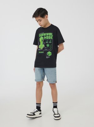 T-shirt in cotone nero con grafica verde di Minecraft e testo "SURVIVAL MODE". Abbinato a pantaloni bassi in denim chiaro e sneakers bianche e nere.