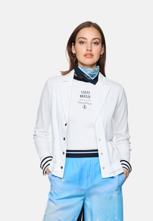 Woman wearing white blazer, white "Salty Breeze" shirt, blue scarf, and light blue shorts with sailboat and seagull print, hands in pockets.