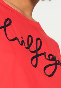 Red cotton T-shirt featuring black embroidered text design with curved lettering. Smooth texture, short sleeves, and rounded neckline.