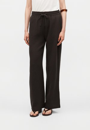 Femme portant un pantalon noir ample texturé avec cordon de serrage, jambes larges et sandales noires à bout ouvert sur un fond uni.