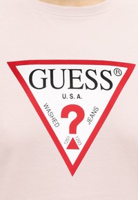 Ερυθρό και λευκό τριγωνικό λογότυπο Guess σε άσπρο ύφασμα, με ένα μεγάλο ερυθρό ερωτηματικό και το κείμενο «GUESS U.S.A. Washed Jeans» μέσα στο τρίγωνο.