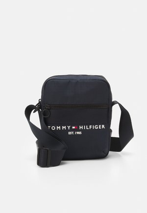 Kompakte schwarze Umhängetasche mit verstellbarem Riemen, vordere Reißverschlusstasche und "Tommy Hilfiger EST. 1985"-Logo mit roter, weißer und blauer Fahne.