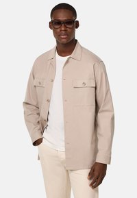 Giacca beige chiaro in cotone, con chiusura a bottoni, due tasche sul petto e maniche lunghe. Indossata sopra una t-shirt bianca e pantaloni crema.