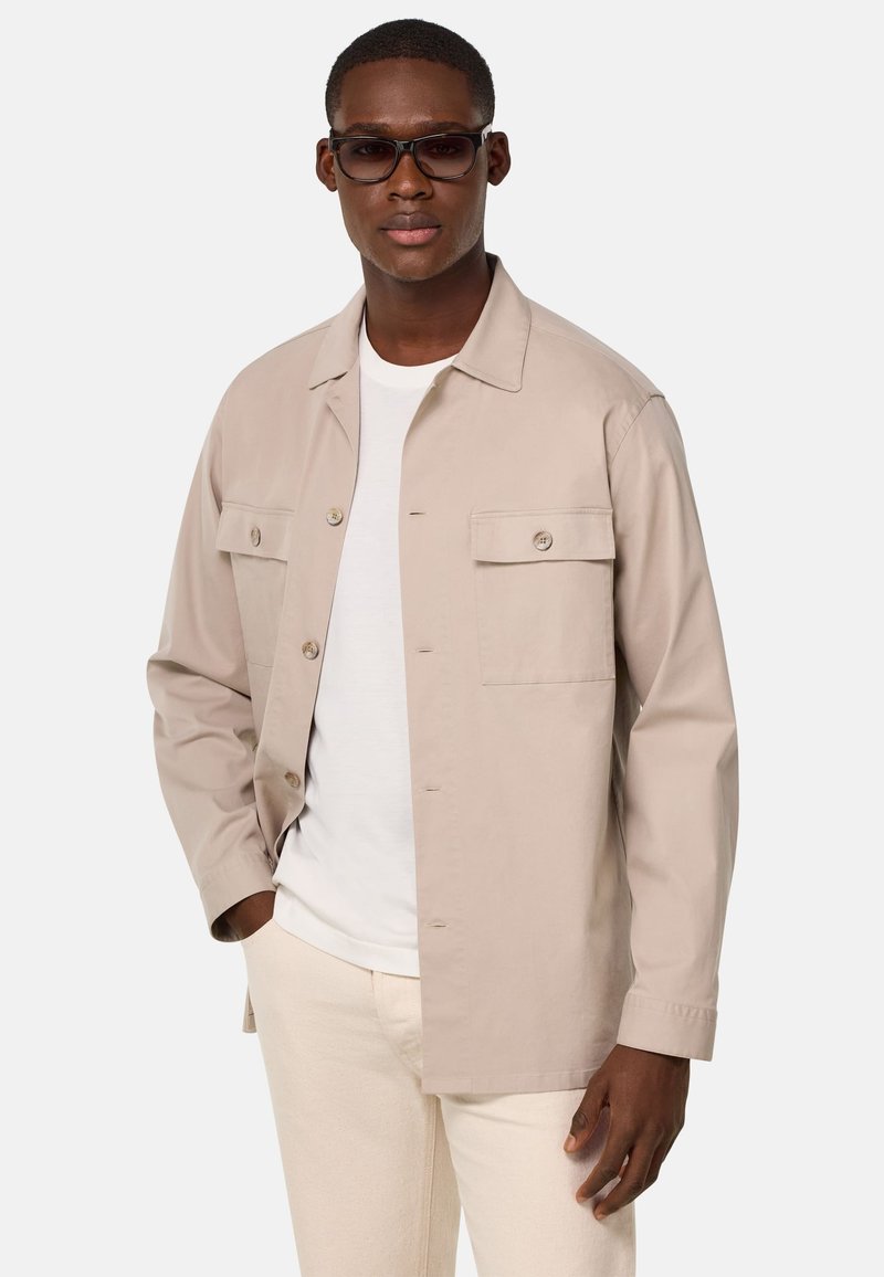 Giacca beige chiaro in cotone, con chiusura a bottoni, due tasche sul petto e maniche lunghe. Indossata sopra una t-shirt bianca e pantaloni crema.