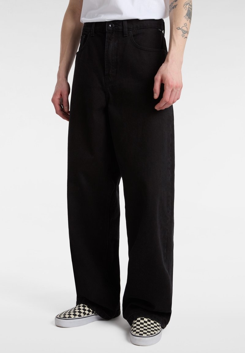 Personne portant un pantalon noir à jambes larges, une chemise blanche et des chaussures slip-on à carreaux noirs et blancs, debout devant un fond uni.