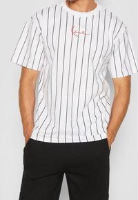 Mężczyzna w białej koszulce z krótkim rękawem w cienkie czarne pionowe paski oraz czerwoną haftowaną naszywką na piersi, w zestawieniu z czarnymi spodniami.