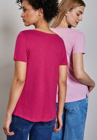 Dos mujeres están de espaldas, llevando camisetas. Una camiseta es fucsia y la otra es rosa claro. Ambas camisetas tienen mangas cortas y un corte holgado.