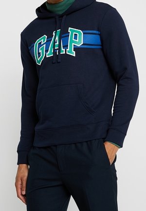 Mand i mørkeblå GAP-hoodie med grønt og hvidt logo samt blå striber, iført mørke bukser, hænder afslappede, den ene i lommen.