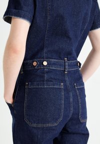 Combinaison en denim foncé à manches courtes, coutures visibles, boutons-pression en cuivre et deux poches arrière. Texture lisse avec une coupe ajustée.