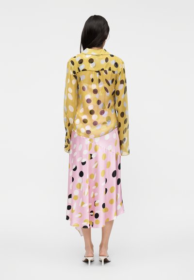 Διαφανές κίτρινο πουκάμισο με μεγάλα πολύχρωμα πουά, συνδυασμένο με ροζ ασύμμετρη φούστα με μαύρα και χρυσά στίγματα. Παπούτσια με τακούνι.