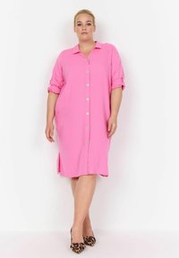 Abito rosa con bottoni, maniche corte, tasche frontali, spacco laterale e vestibilità comoda. Abbinato a tacchi con stampa leopardata. Tessuto dalla texture liscia.