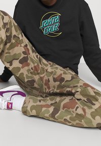 Black sweatshirt with embroidered "SANTA CRUZ" logo, paired with tan camo pants. Footwear features purple accents and a white base.