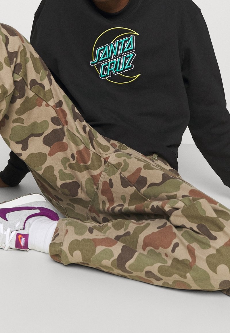 Black sweatshirt with embroidered "SANTA CRUZ" logo, paired with tan camo pants. Footwear features purple accents and a white base.