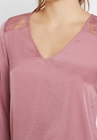 V-pääntie, pitkähihainen paita pehmeästä mauven värisestä kankaasta, jossa pitsejä olkapäillä, omaten sileän pinnan ja rentoa istuvuutta.