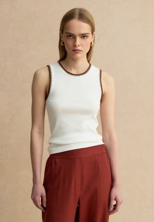 Joven mujer con cabello rubio lacio, lleva una blusa blanca sin mangas con ribete marrón y pantalones de talle alto color óxido, sobre un fondo beige.