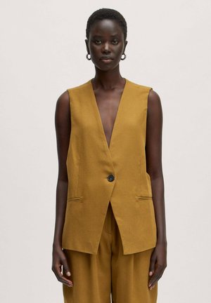 Vrouw met kort haar die een mosterdgele mouwloze blazer met één knoop en bijpassende losse broek draagt tegen een effen achtergrond.
