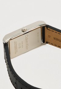 Montre rectangulaire en acier inoxydable Calvin Klein avec un bracelet en cuir noir. Le dos gravé affiche le logo de la marque et des détails techniques.