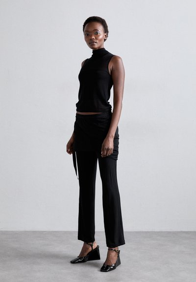 Top preto sem mangas com gola alta, combinado com saia envolvente preta e calças justas. Sapatilhas pretas de verniz com cunha completam o visual.