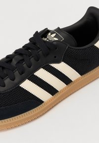 Svarta textilsneakers med läderdetaljer, featuring tre krämfärgade ränder, en strukturerad ovansida och en gummigummisula.