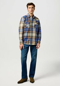 Camisa de cuadros de manga larga en tonos azul, beige y marrón, con dos bolsillos en el pecho y cuello abotonado, combinada con jeans azules y botas marrones.