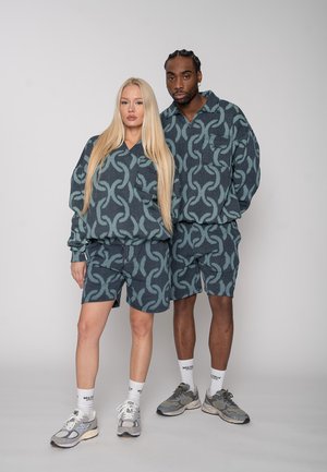 Zwei Erwachsene tragen passende gemusterte langärmlige Oberteile und Shorts in Marineblau und Hellblau und stehen selbstbewusst vor einem einfarbigen Hintergrund.