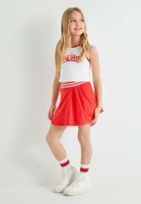 Hvid crop top med røde kanter og teksten "CLARKS", parret med en rød flared nederdel. Båret med hvide sneakers og sokker med røde accenter.