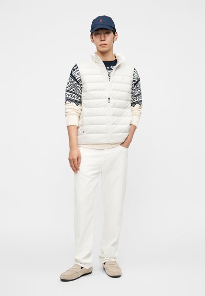 Επενδυμένο λευκό γιλέκο πάνω από μπλε πουλόβερ με σχέδιο, συνδυασμένο με παντελόνι σε κρεμ απόχρωση και μπεζ παπούτσια τύπου slip-on· μπλε καπέλο του μπέιζμπολ με λογότυπο.