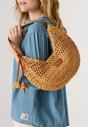 Žena v modrom pruhovanom tričku nesie na ramene béžovú tkanú polmesiacovú kabelku s koženým remienkom a strapcovými detailmi.