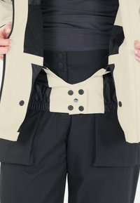 Hellebeige Jacke mit schwarzen Akzenten, ausgestattet mit einem Reißverschluss, Druckknöpfen und einem strukturierten Futter. Schwarze Hose mit Seitentaschen.