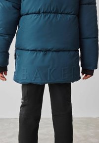 Veste doudoune bleue avec texture matelassée et logo "BAKER" en relief au bas. Portée sur un pantalon noir, mettant en valeur une coupe décontractée.