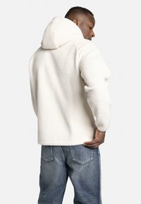 Witte fleece hoodie met een textuur, voorzien van een capuchon, gecombineerd met blauwe denim jeans, gezien vanaf de achterkant.