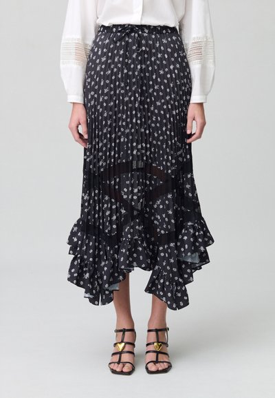 Jupe plissée noire avec impression florale blanche, ourlet volé et taille à nouer. Portée avec des sandales à lanières noires ornées d'accents en forme de cœur dorés.