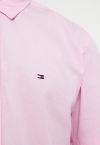 Ανοιχτό ροζ πουκάμισο με γιακά και μικρό ορθογώνιο λογότυπο κεντημένο στο αριστερό στήθος, με χρώματα ναυτικό μπλε, άσπρο και κόκκινο.