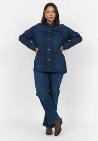 Denim jakna in hlače v temno modri barvi, s vidnimi šivi, dvema sprednjima žepoma in srebrnimi gumbi. Obute v rjave čevlje s peto.