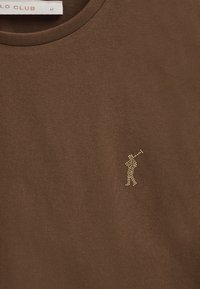 Camiseta de manga corta marrón hecha de una tela suave. Presenta un pequeño logo de golfista con tachuelas doradas en el área del pecho izquierdo. Cuello redondo.