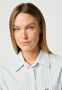 Camicia a righe blu chiaro realizzata in tessuto morbido, con colletto, apertura a bottoni e una piccola etichetta logo sulla tasca sinistra del petto.