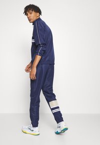 Navy blue tracksuit with white stripes, zip-up jacket, elastic cuffs, matching pants, and grey accent on the lower leg. White sporty sneakers.