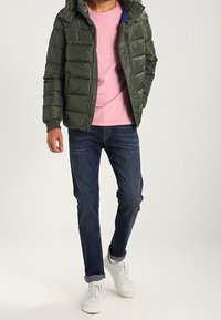 Olivgrön pufferjacka med dragkedja fram, rosa t-shirt under, mörka jeans och vita sneakers. Jackan har huva och horisontell sömnad.