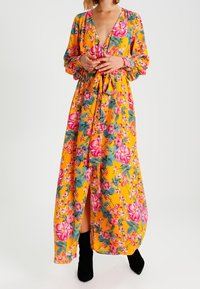 Maxiklänning i starkt gult med blommönster som har rosa och gröna accenter. Långa ärmar, djup V-ringning och midjebandsdetalj.
