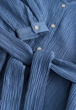 Blue textured fabric shirt featuring vertical pleats, button fastenings, and a matching waist tie. Noticeable light-colored buttons.