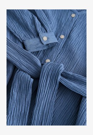 Blå tekstureret stofskjorte med lodrette folder, knaplukninger og et matchende taljebånd. Bemærkelsesværdige lysfarvede knapper.